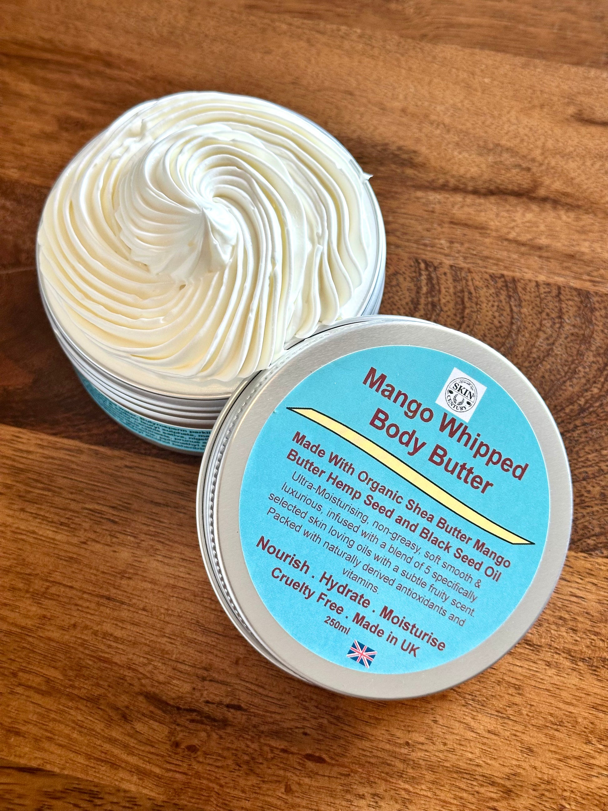 An open round tin of Mango Whipped Body Butter showing thick whipped cream-colored butter, with the lid placed beside it on a wooden surface. The lid label is light blue with dark red and yellow text.